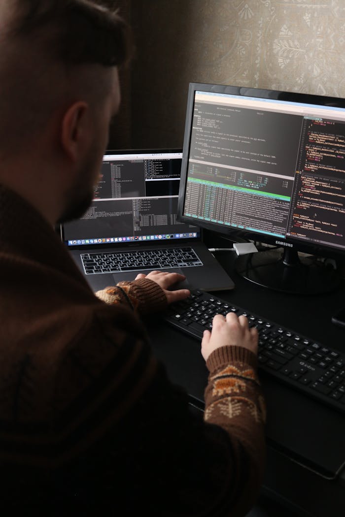 A programmer working on code with dual monitors and a laptop in a dimly lit room.